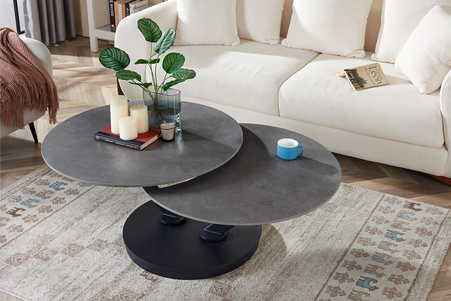 Een moderne woonkamer met een crèmekleurige bank, een vloerkleed met patroon en twee overlappende Salontafel Lichtgrijs, Roest Rond Steen. Op de tafels staan kaarsen, een plant, boeken en een blauw koffiekopje. Op de bank ligt een boek.