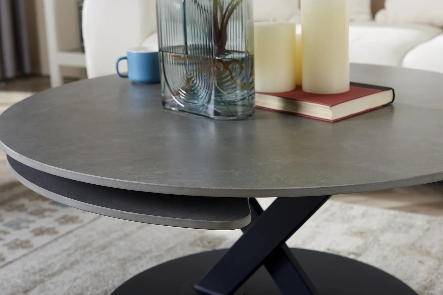 De Salontafel Lichtgrijs, Roest Rond Steen toont een boek, twee pilaarkaarsen, een blauwe mok en een glazen vaas met plantenstengels. Op de achtergrond staan een crèmekleurige bank en een vloerkleed met patroon.