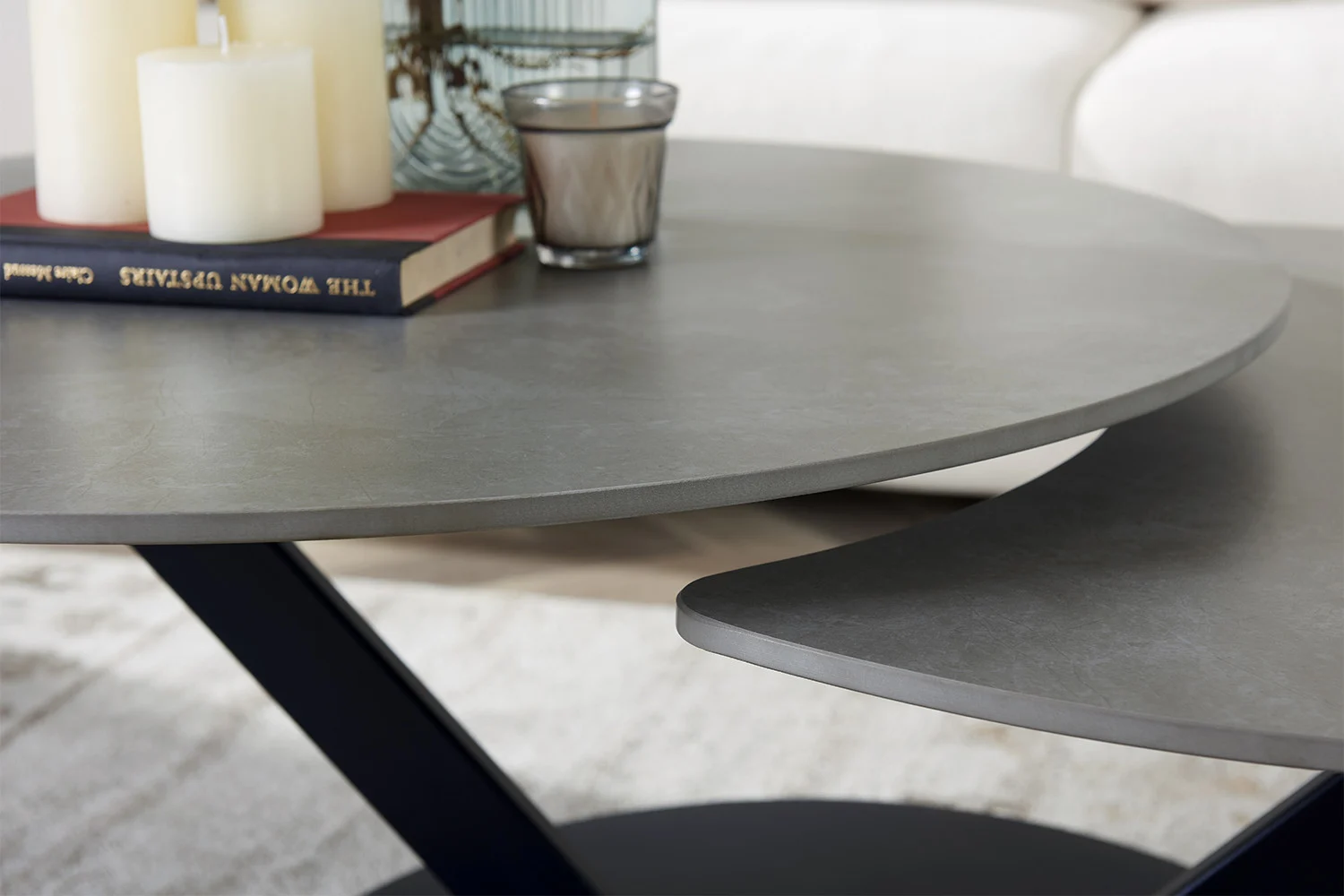 Een close-up van de Salontafel Lichtgrijs, Roest Rond Steen met gelaagde oppervlakken. Bovenop staan drie pilaarkaarsen, een glazen kaarsenhouder en gestapelde boeken. Op de achtergrond staan een beige bank en vloerkleed.