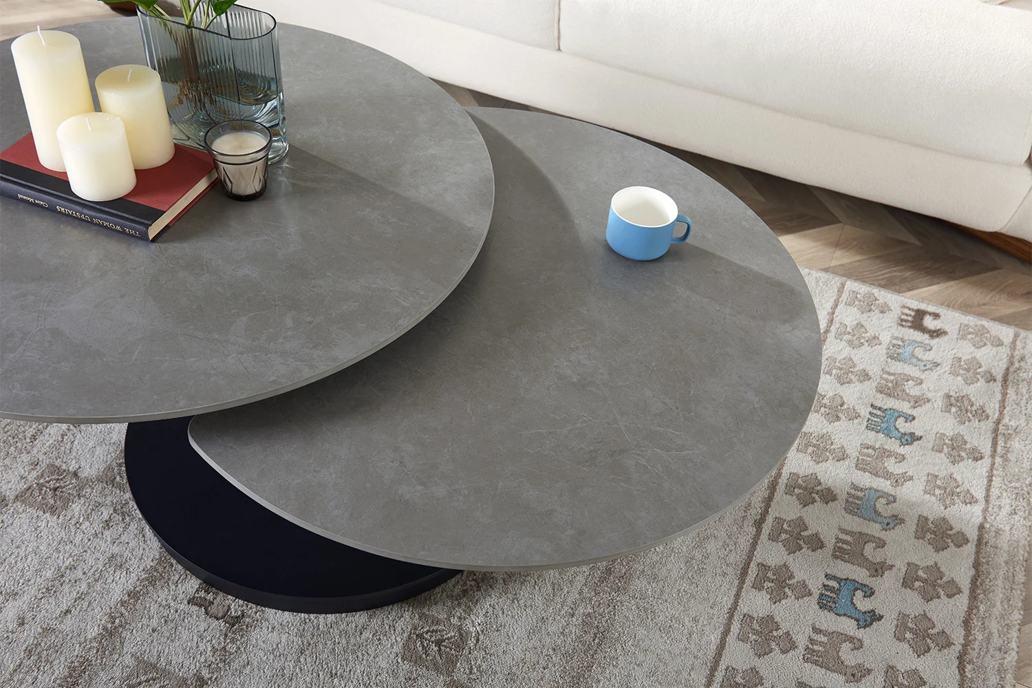 De Salontafel Lichtgrijs, Roest Rond Steen, een moderne ronde tafel met twee niveaus, toont kaarsen, een boek, een glazen vaas en een blauwe beker. Het staat op een licht vloerkleed naast een beige bank en een gedessineerde mat met blauwe en bruine motieven.