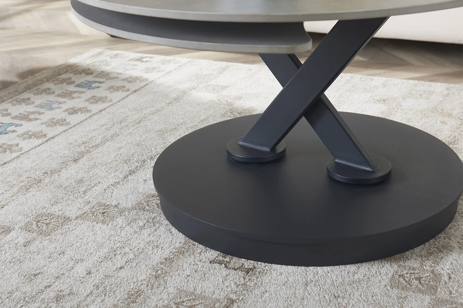 Close-up van de Salontafel Lichtgrijs, Roest Rond Steen met een zwart X-vormig metalen onderstel, geplaatst op een beige en lichtbruin gedessineerd tapijt op een houten vloer.