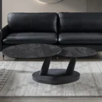 Een moderne woonkamer met een zwarte leren bank, twee Salontafel Donkergrijs, Roest Rond Steen salontafels en een vloerkleed met patroon op een lichte houten vloer.