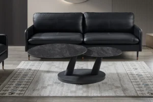 Een moderne woonkamer met een zwarte leren bank, twee Salontafel Donkergrijs, Roest Rond Steen salontafels en een vloerkleed met patroon op een lichte houten vloer.