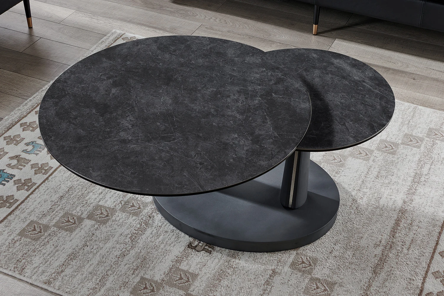 Een moderne, ronde salontafel Donkergrijs, Roest Rond Steen met twee overlappende ronde etages staat op een vloerkleed met patroon op een lichte houten vloer.