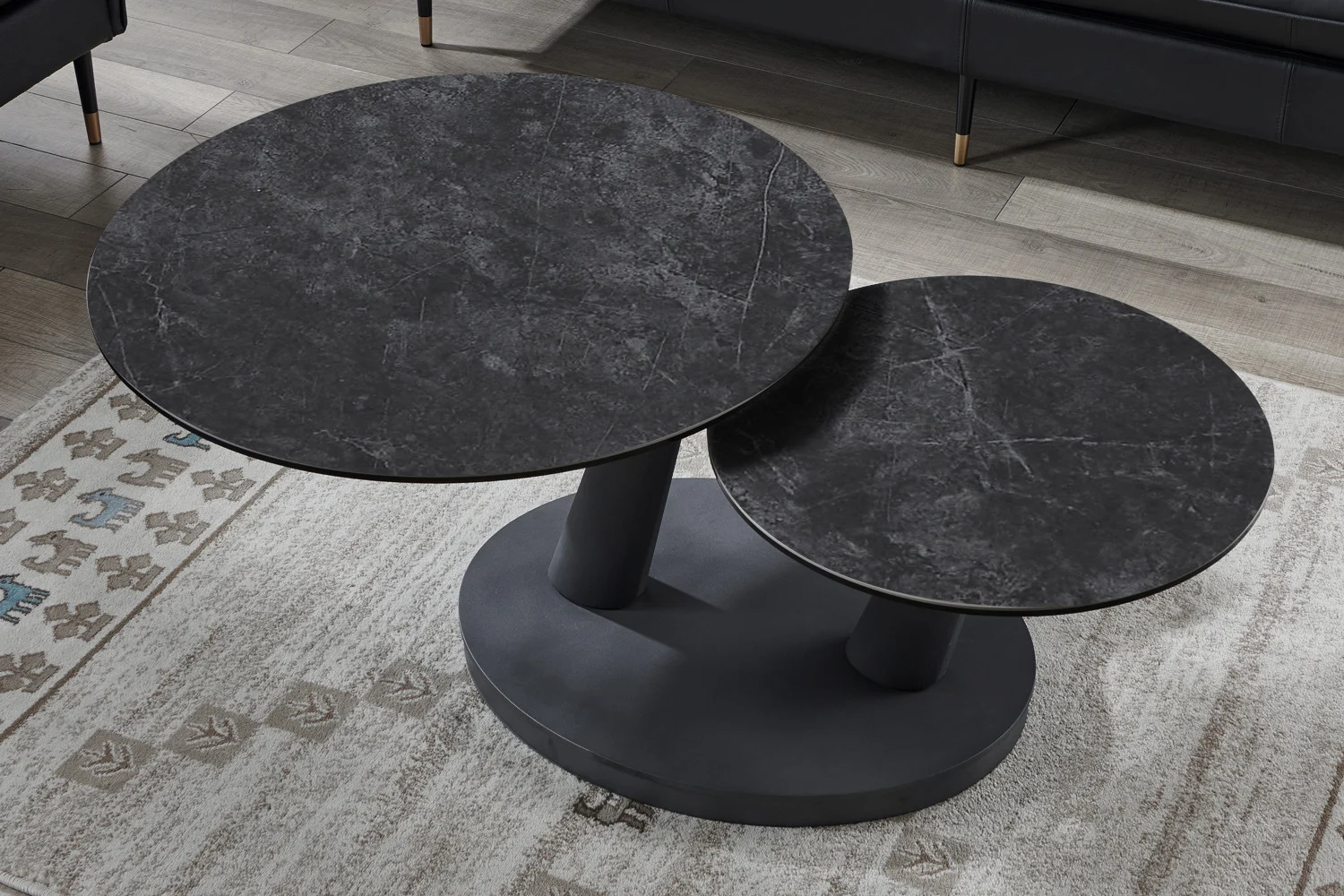 De Salontafel Donkergrijs, Roest Rond Steen heeft twee ronde, donkere stenen bladen op verschillende hoogtes met cilindrische poten op een ovale basis, gezien op een tapijt met patroon en een houten vloer.