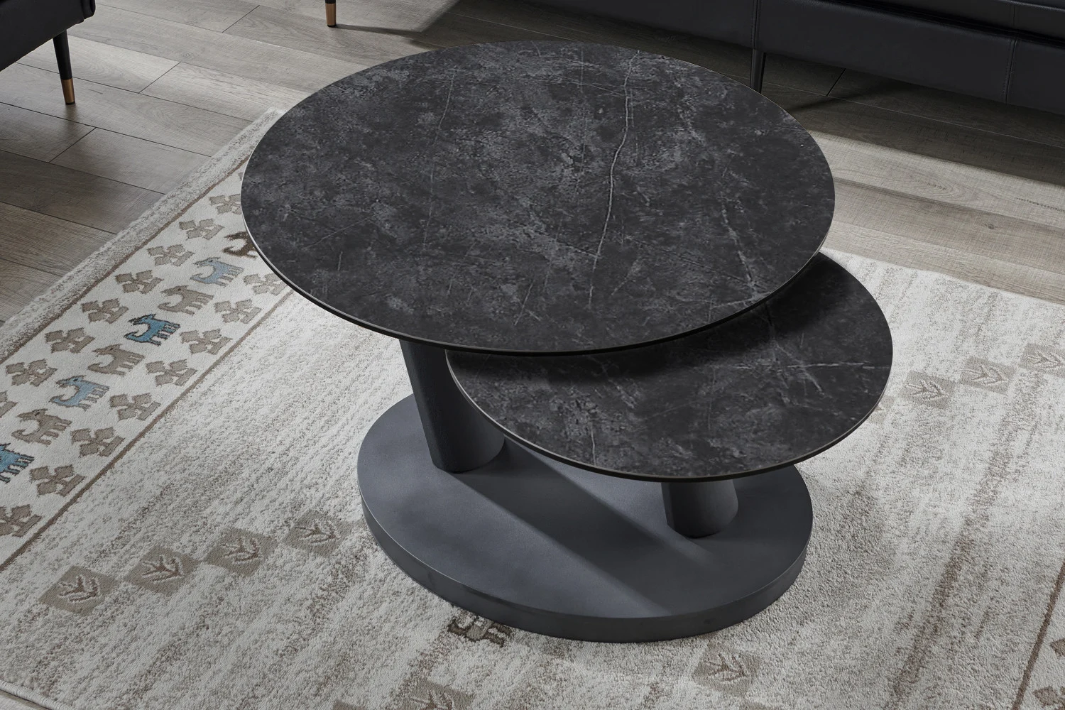 De Salontafel Donkergrijs, Roest Rond Steen - een moderne, ronde, donkergrijze stenen salontafel met twee niveaus - staat op een beige geometrisch tapijt op een houten vloer bij een zwarte bank.