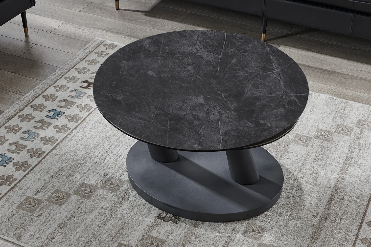 De Salontafel Donkergrijs, Roest Rond Steen heeft een rond donkergrijs stenen blad en een moderne sokkel, geplaatst op een licht gedessineerd tapijt op een houten vloer met donkere banken zichtbaar op de achtergrond.