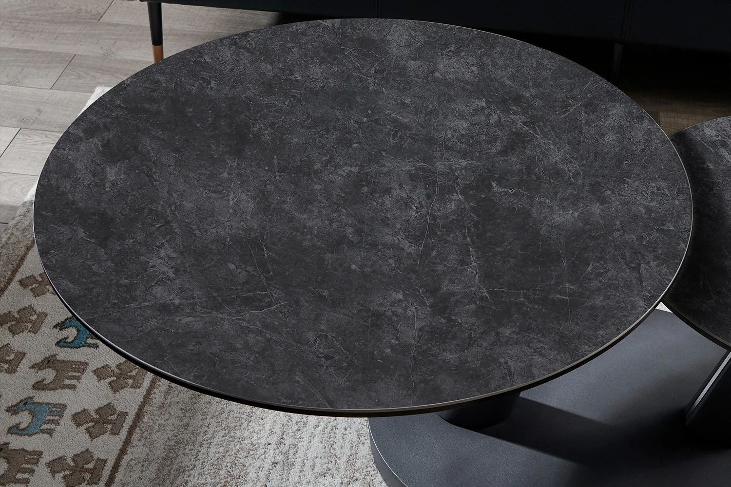 De Salontafel Donkergrijs, Roest Rond Steen is een ronde, donkergrijze stenen salontafel met subtiele witte adering, geplaatst op een vloerkleed met dierenmotieven naast een houten vloer.