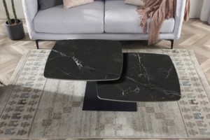 Een moderne woonkamer met een lichtgrijze bank, een beige vloerkleed met patroon en de Salontafel Zwart Rond Steen - een ronde zwarte stenen salontafel - geplaatst naast een potplant en geaccentueerd met een roze deken.