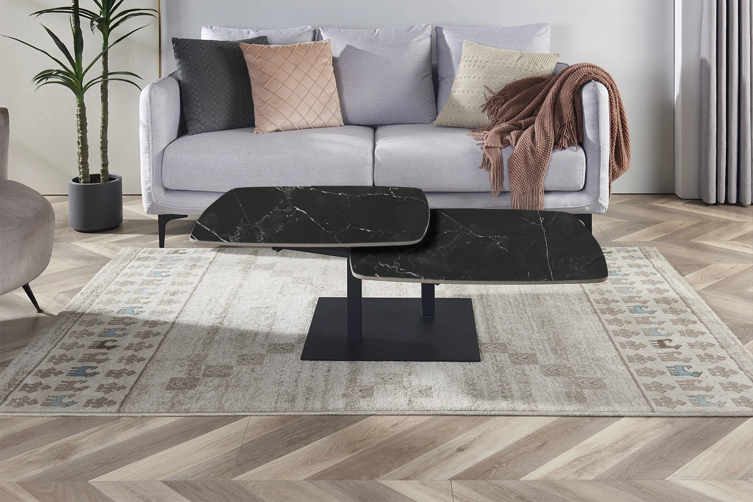 Een moderne woonkamer met een lichtgrijze bank, roze en beige kussens, een deken, de Salontafel Zwart Rond Steen, een vloerkleed met patroon, een plant en een houten vloer met chevronpatroon.