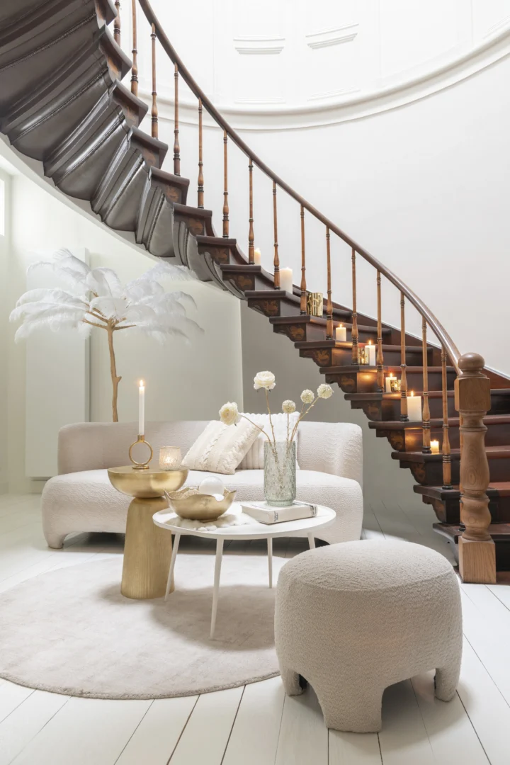 Een lichte, moderne leefruimte met de Bijzettafel Wit 38 CM Aluminium, een gebogen houten trap, witte en beige meubels, ronde tafels, kaarsen langs de trap en een decoratief wit verenarrangement in zachte, gezellige neutrale tinten.