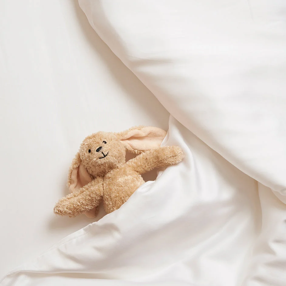 Zachte bruine knuffelkonijn in een wit TENCEL™ hoeslaken. Boven aanzicht, zachte belichting, gladde stof, kinderkamer, rustgevend.