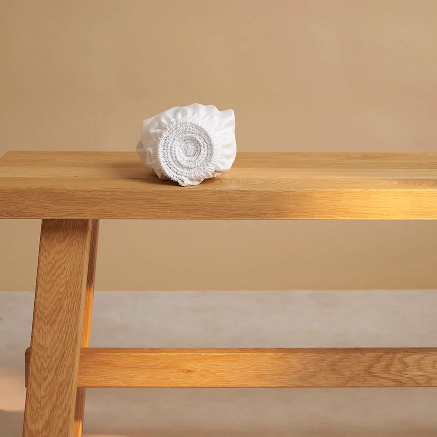 Opgerold wit hoeslaken van TENCEL™ sateen op houten bank. Zachte belichting, beige achtergrond. Textuur van het materiaal is goed zichtbaar.