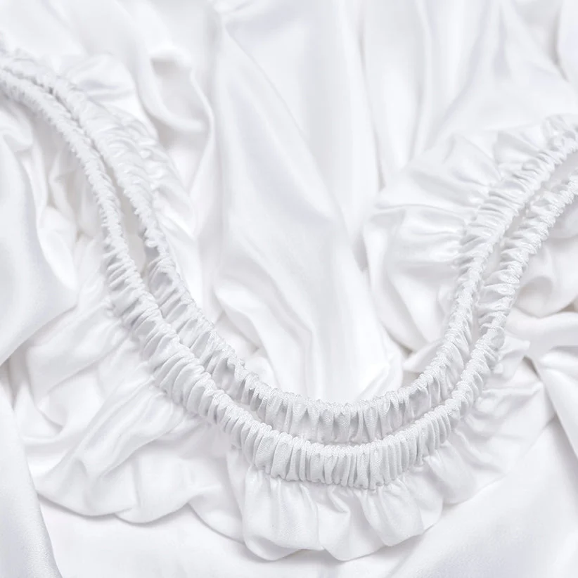 Detailopname van een wit hoeslaken met elastiek. De stof is zacht en glanzend, vermoedelijk TENCEL™ sateen, en ligt in plooien. Heldere belichting.