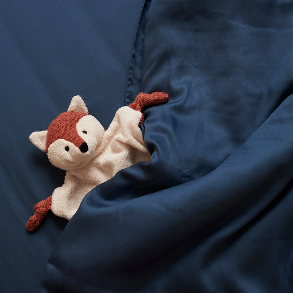 Van bovenaf: blauw TENCEL™ hoeslaken met zachte glans, met een knuffelvos. De stof is glad en valt in plooien, zacht licht.