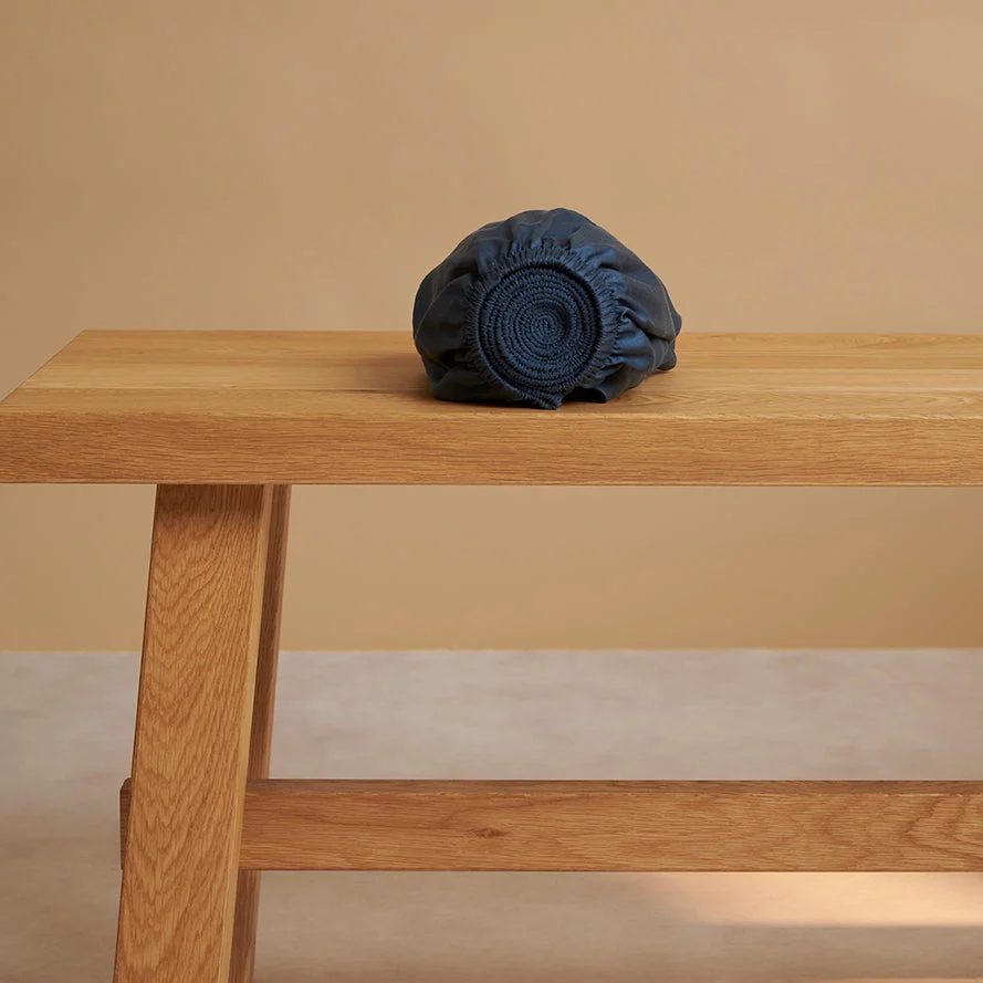 Oprolbaar blauw hoeslaken van TENCEL™ Sateen op een houten bank. Zachte belichting, warme achtergrondkleur. Hypoallergeen en comfortabel.