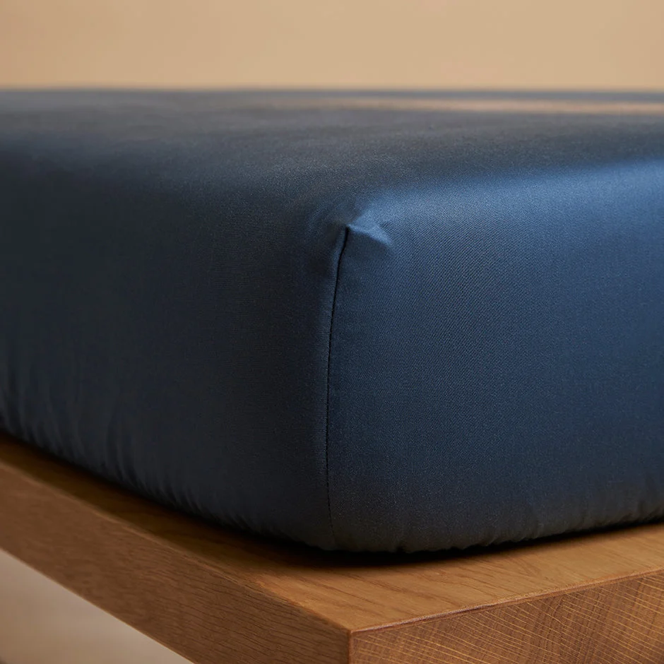 Detailopname van een blauw hoeslaken van TENCEL™ Lyocell op een houten matras. Zacht licht accentueert de gladde textuur.