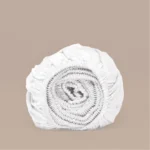 Opgerold wit hoeslaken van TENCEL Lyocell, zacht satijn. Elastiek is zichtbaar. Neutrale achtergrond, zachte belichting.