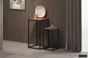 Een moderne hoek met bijzettafel 'Madeira' 2-delig Mango hout-twee geneste vierkante tafels, zwart metalen frames, mangohouten bladen. Op de langste staan een bord en een vaas, op de kortere een abstract beeldhouwwerk. Grijze gordijnen en muurkunst maken de ruimte compleet.