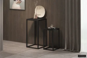 De Bijzettafel 'Madeira' 2-delig Mango hout zwart, twee zwarte mangohouten bijzettafels van verschillende hoogtes, staan bij een wand met houten panelen. Ze tonen decoratieve voorwerpen, met een ingelijst portret en lange grijze gordijnen in de buurt.