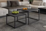 Een Salontafel Zwart Mangohout Compact staat op een tapijt met grijs patroon, met daarop een schaal met groene appels en peren, terwijl een andere een glazen vaas met een kaars en wat sinaasappels vasthoudt. Een beige sofa verschijnt op de achtergrond.