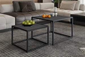Een Salontafel Zwart Mangohout Compact staat op een tapijt met grijs patroon, met daarop een schaal met groene appels en peren, terwijl een andere een glazen vaas met een kaars en wat sinaasappels vasthoudt. Een beige sofa verschijnt op de achtergrond.