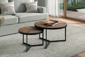 De Salontafel Mangohout Compact, een ronde houten salontafel met zwarte metalen poten, staat op een lichtgrijs vloerkleed in een moderne woonkamer met een grijze bank en een groot raam. De ene tafel biedt plaats aan een tijdschrift en is iets hoger dan de andere.
