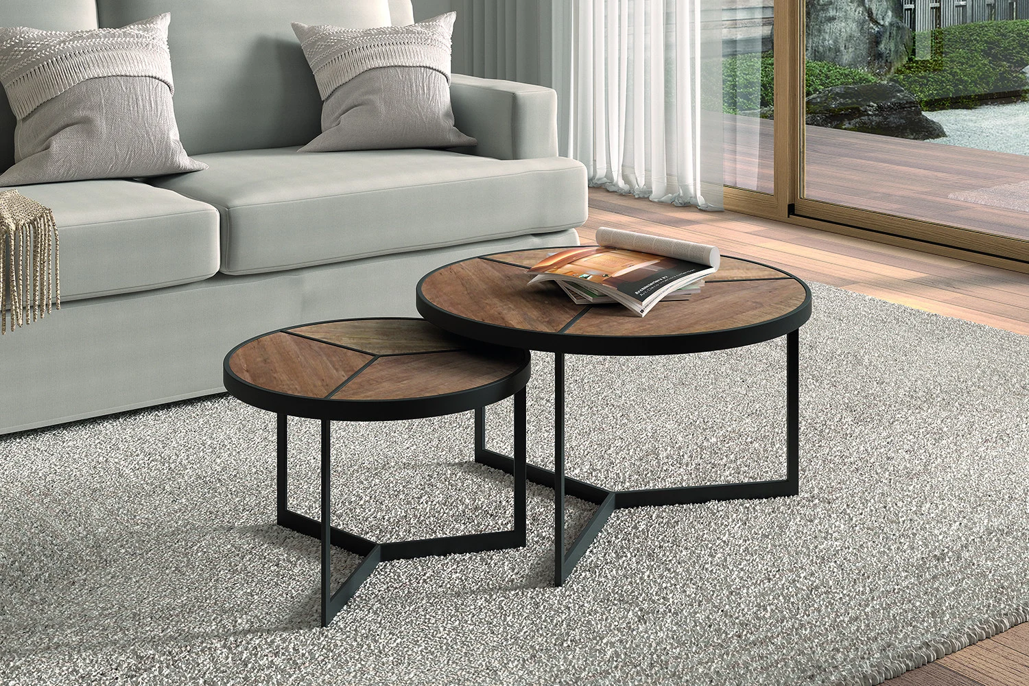De Salontafel Mangohout Compact, een ronde houten salontafel met zwarte metalen poten, staat op een lichtgrijs vloerkleed in een moderne woonkamer met een grijze bank en een groot raam. De ene tafel biedt plaats aan een tijdschrift en is iets hoger dan de andere.