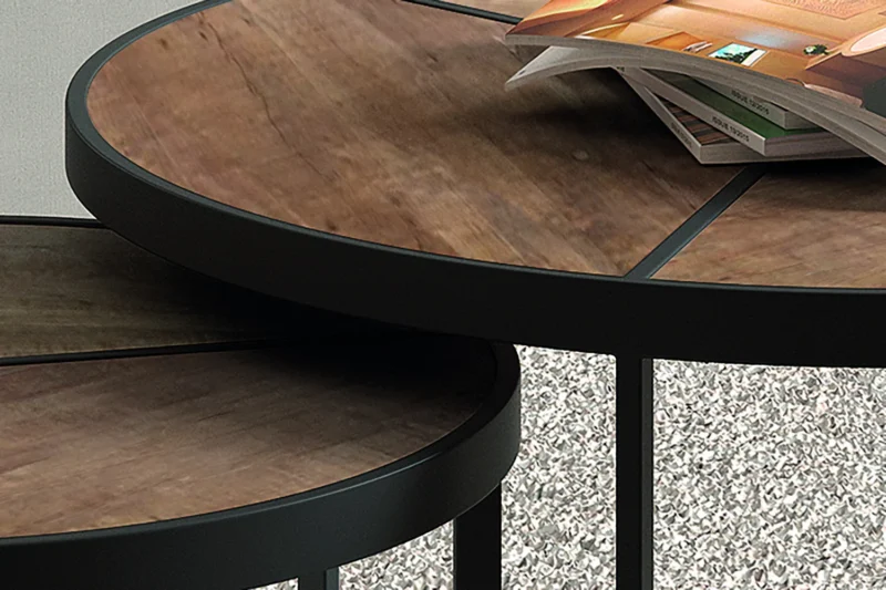Een close-up van twee Salontafel Mangohout Compact nesttafels met donkere metalen frames en houten bladen. Op de grotere tafel ligt een tijdschrift en beide staan op een tapijt met structuur.