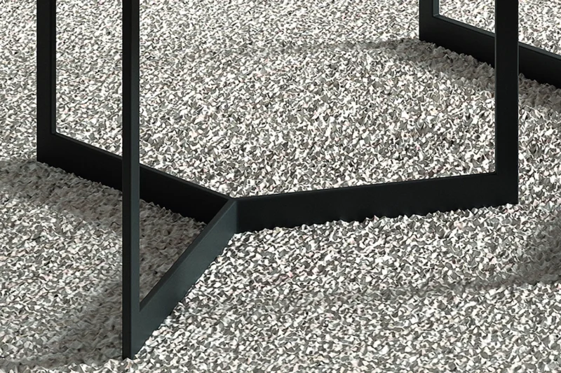 Close-up van de zwarte metalen poten van Salontafel Mangohout Compact die geometrische vormen vormen op een gestructureerd, lichtgrijs grindoppervlak.