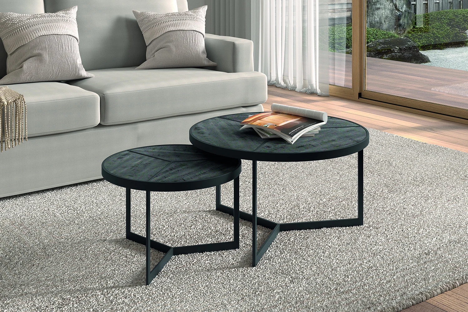 Twee ronde Salontafel Zwart Mangohout Design salontafels met metalen poten, voorzien van een tijdloos ontwerp, staan op een lichtgrijs tapijt bij een beige bank. Een open tijdschrift rust op de grotere tafel, met grote ramen die een houten dek buiten tonen.