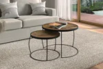 Salontafel Mangohout Industrieel Tijdloos toont twee ronde nesttafels met houten bladen en zwarte metalen frames, geplaatst op een beige vloerkleed in een woonkamer met een lichtgrijze bank, gezellige kussens, een deken en grote ramen die uitkijken op de tuin.
