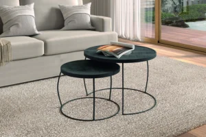 De Salontafel Zwart Mangohout Design Modern, een set ronde zwarte salontafels met ranke metalen poten, staat op een beige tapijt in een moderne woonkamer met een bank met kussens, transparante gordijnen en ramen met uitzicht op de tuin.