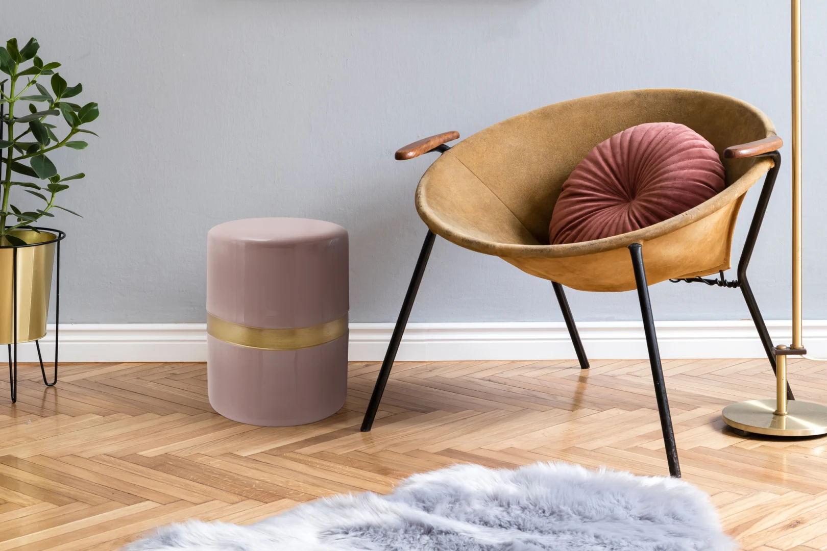 Een gezellige moderne kamer is voorzien van een ronde bruine stoel met een roze geplooid kussen, de Bijzettafel Roze Rond Ijzer Compact met een gouden band, een plant in een gouden pot en een donzig grijs vloerkleed op een visgraat houten vloer.
