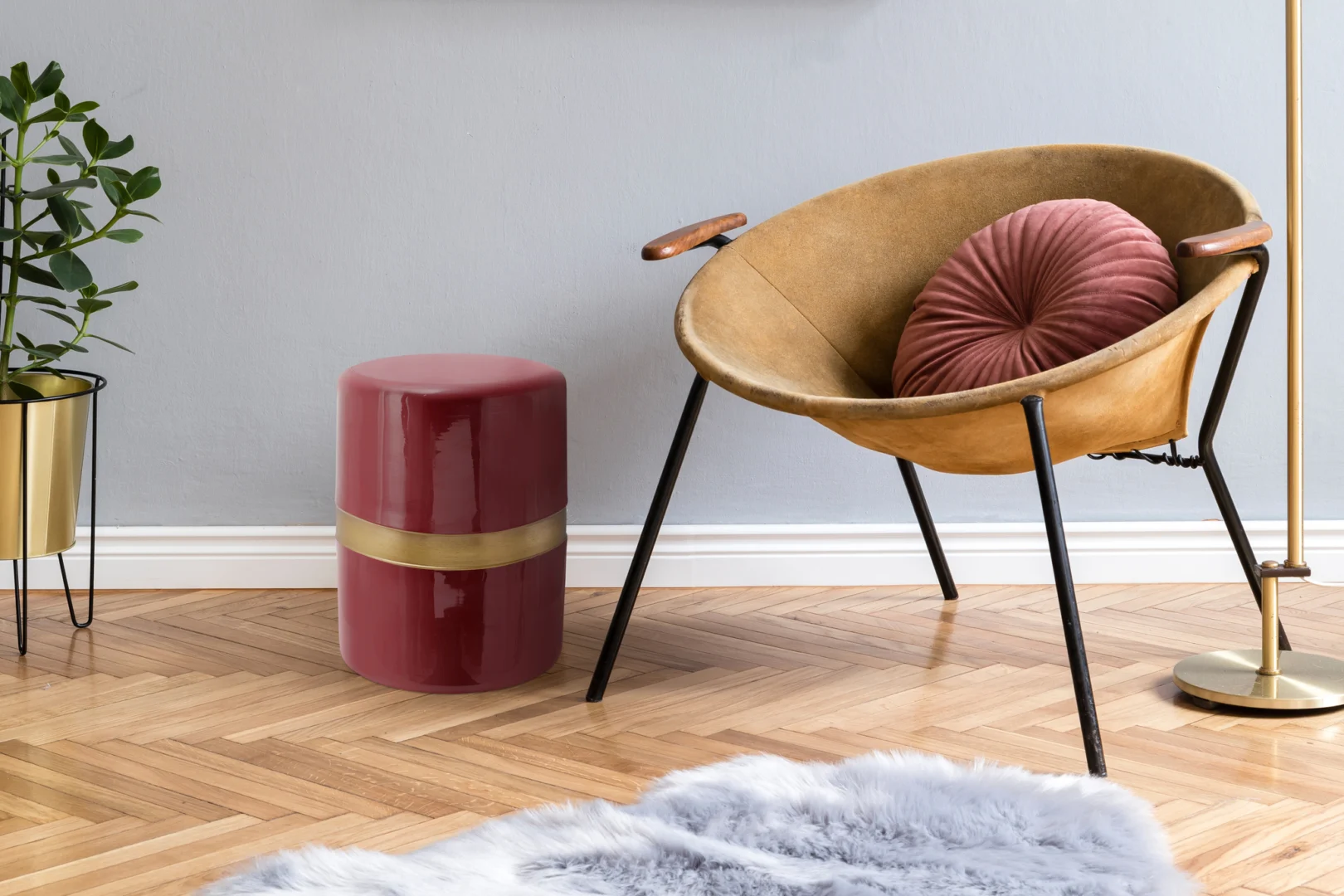 Een moderne woonkamer is voorzien van een bruine stoel met zwarte poten en een roze rond kussen, een Bijzettafel Rond Ijzer Chique glanzend roze bijzettafel met gouden band, een potplant, een gouden staande lamp en een vloerkleed van grijs imitatiebont op een vloer van visgraathout.