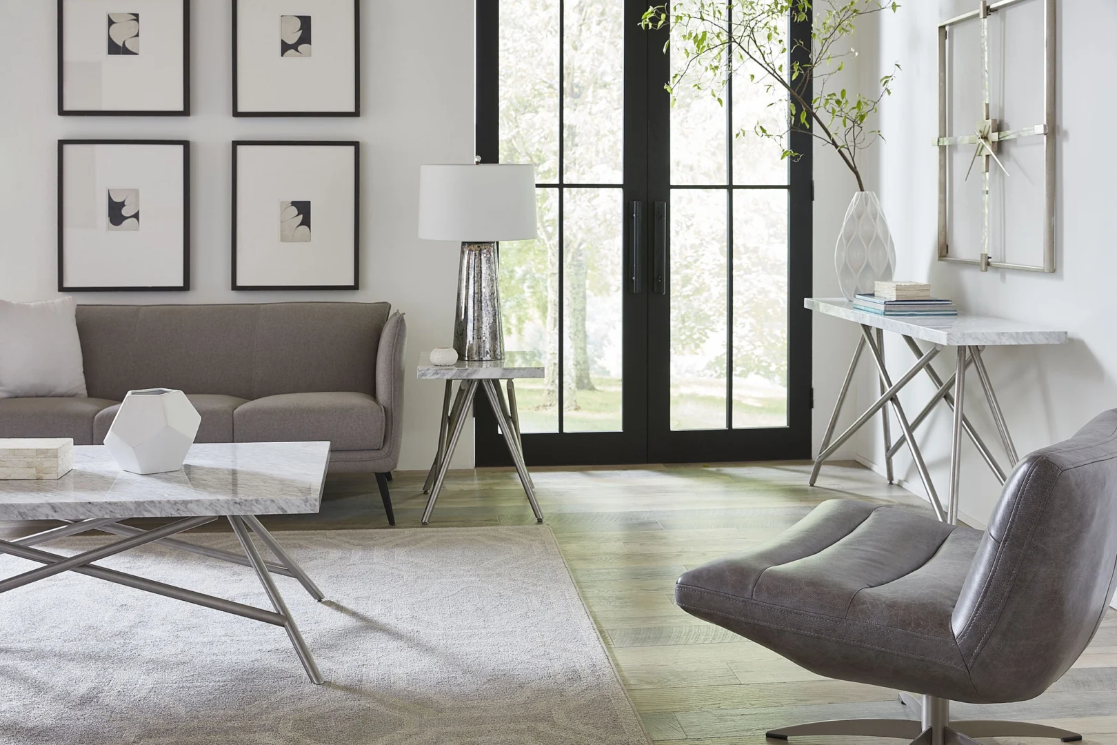 Moderne woonkamer met een grijze bank en stoel, salontafel rvs met marmeren blad en metalen poten, een tafellamp, abstracte muurkunst en hoge glazen deuren die openslaan naar een helder uitzicht naar buiten. Minimalistische inrichting met lichte muren en vloeren.