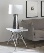 Een moderne woonkamer heeft een grijze bank, de Koraal bijzettafel in marmer met metalen poten, een metalen lamp met een witte kap, een klein decoratief item en twee ingelijste abstracte kunstwerken aan de muur.