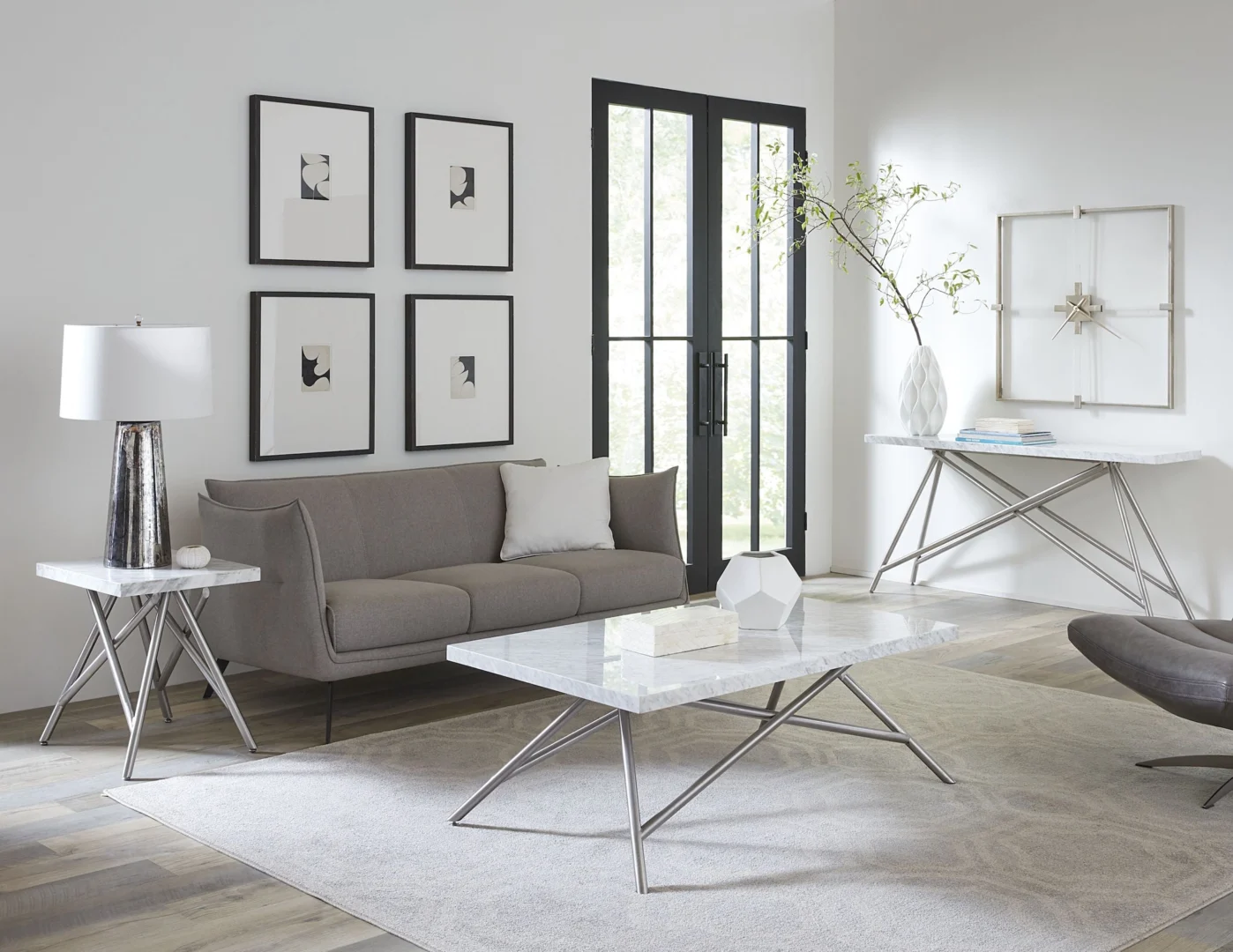 Een moderne woonkamer met een grijze bank, abstracte muurkunst, een decoratieve tak in een witte vaas en grote glazen deuren. De Koraal bijzettafel in marmer voegt elegantie toe, terwijl de neutrale, minimalistische inrichting een schoon, luchtig gevoel creëert.