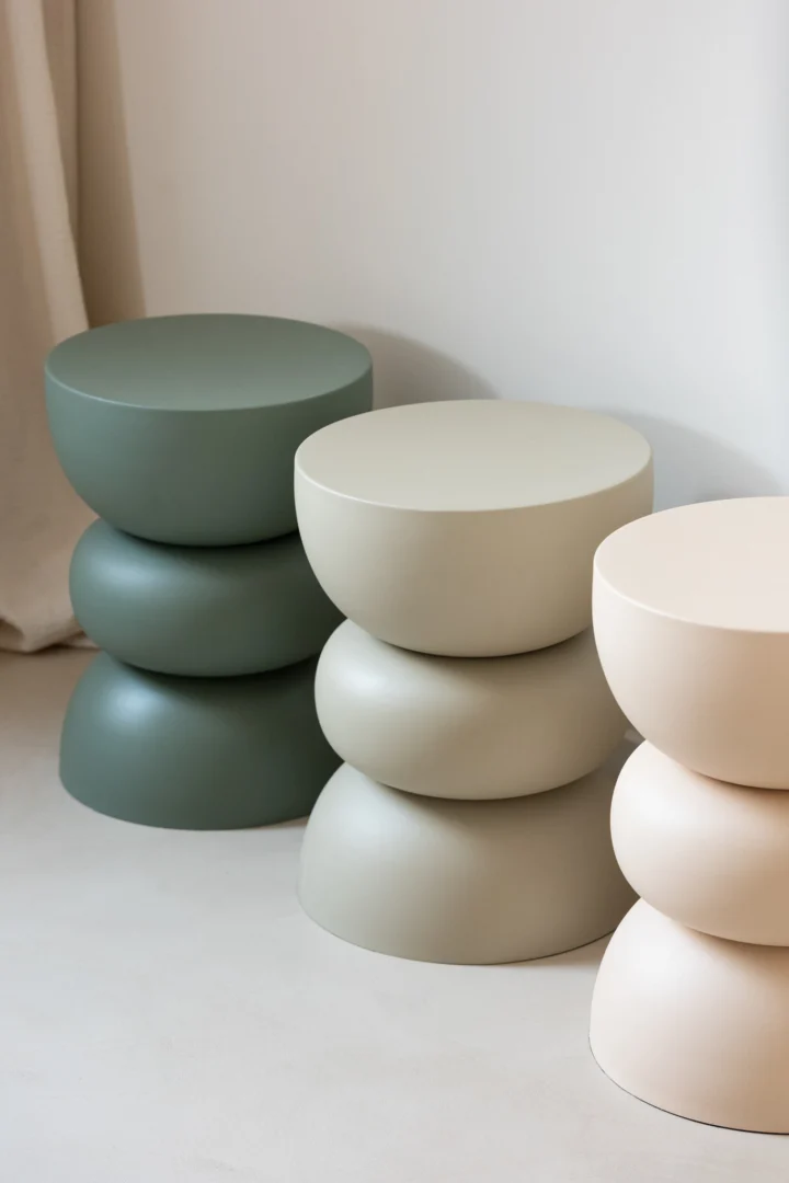 De Bijzettafel Lise IJzer Groen staat samen met twee soortgelijke sculpturale krukjes in lichtgrijs en beige op een rij tegen een neutrale muur. Elk heeft een gestapeld, rond ontwerp en een gladde matte afwerking.
