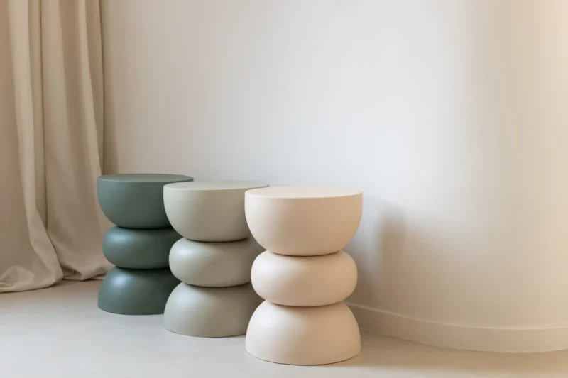 De Bijzettafel Lise IJzer Groen, een moderne bijzettafel in gedempt groen, staat bij een effen witte muur met een licht gordijn aan de linkerkant en voegt een minimalistisch en rustig accent toe aan de ruimte.
