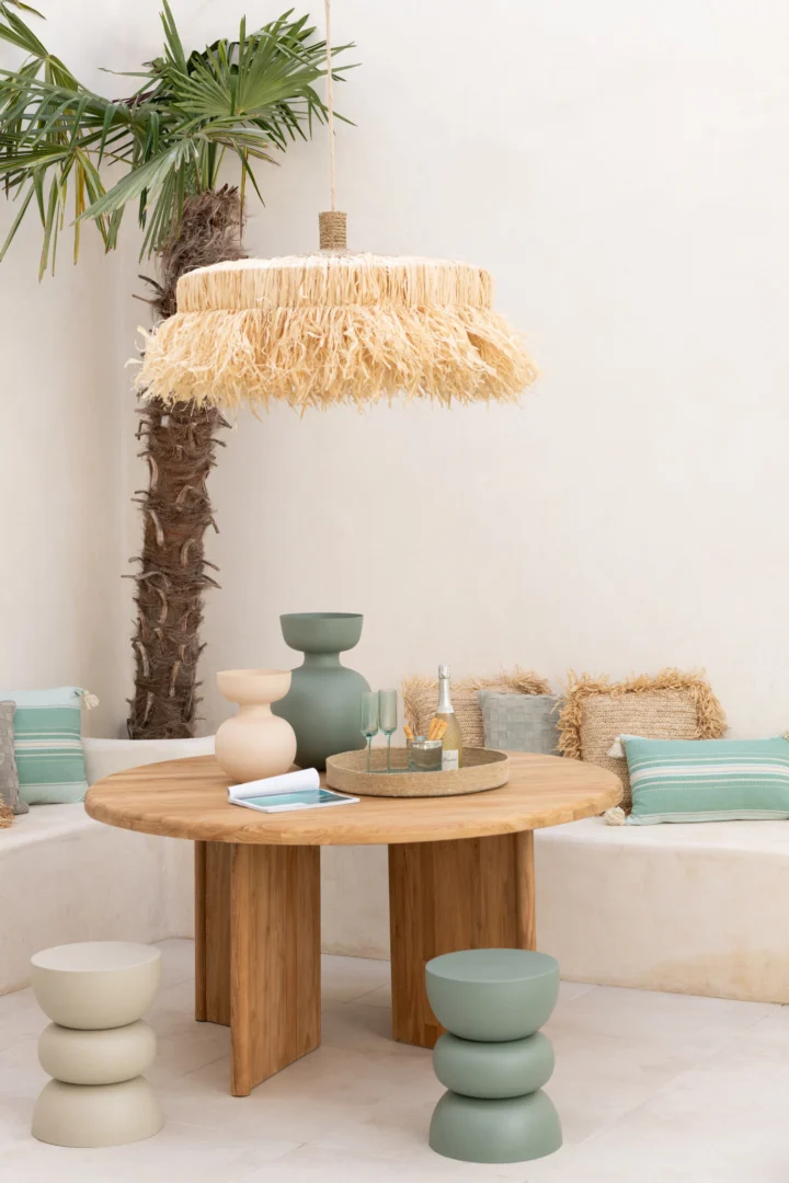De Bijzettafel Lise IJzer Groen zit onder een hanglamp van gefranjerd stro, omringd door banken met kussens en pastelkleurige krukjes. Decoratieve vazen, een dienblad, gestructureerde kussens en een palmboom voegen charme toe aan het tafereel.