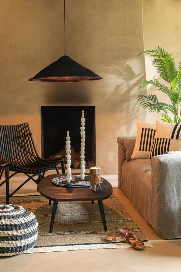 Een gezellige woonkamer met een grijze bank, gestreepte kussens, een zwarte stoel, een rechthoekige salontafel van zwart metaal en teakhout (Salontafel Zwart, Teak 70 x 30 CM), sierkaarsen, een geweven voetenbank en vloerkleed, een plant in pot, sandalen op de vloer en een zwarte hanglamp.