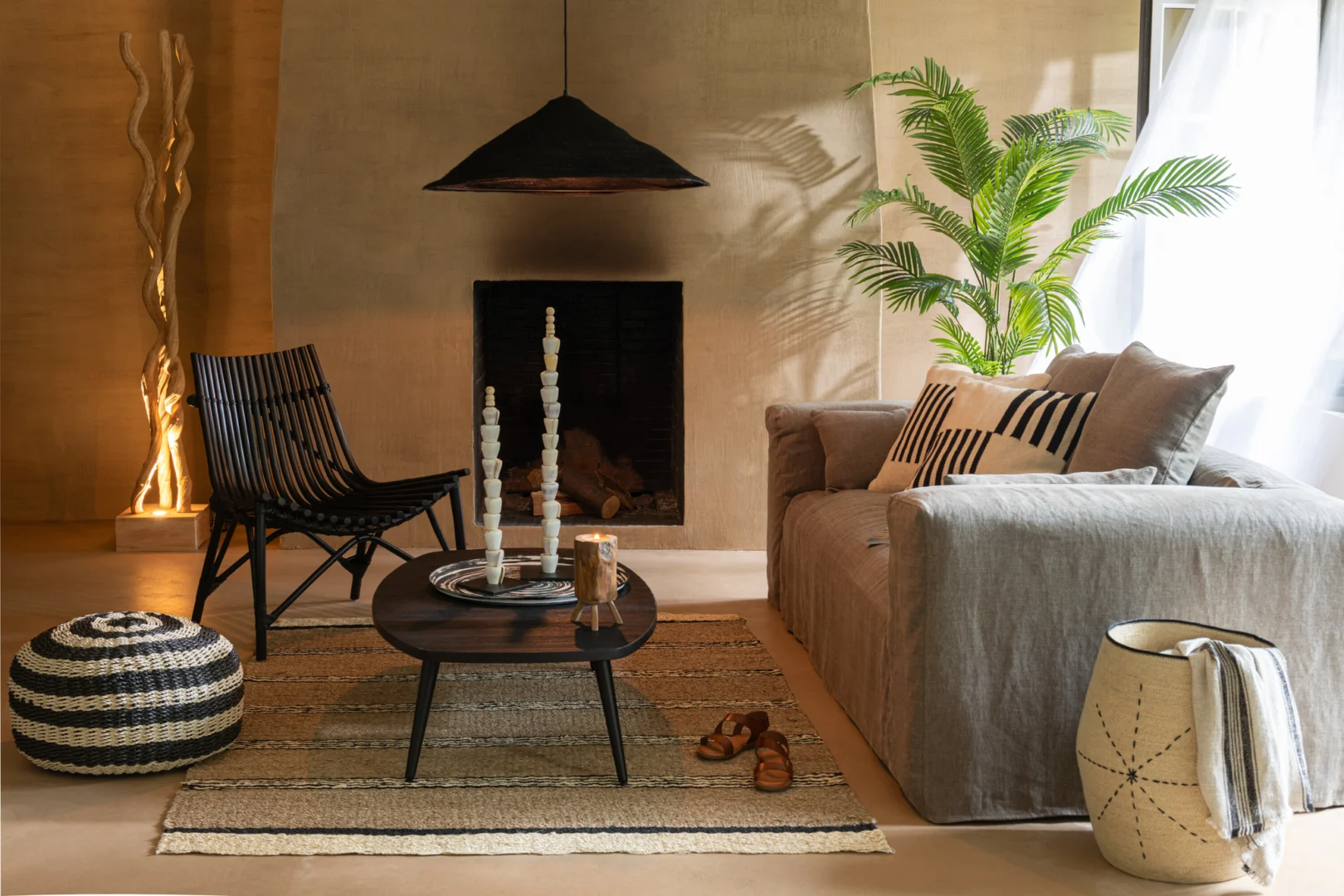 Een gezellige woonkamer met een beige bank, zwarte stoel, gestreepte poef, kamerplant en decoratieve verlichting bij de open haard, met binnenvallend zonlicht en een Salontafel Zwart, Teak 70 x 30 CM - rechthoekig met metalen accenten - in plaats van de salontafel.