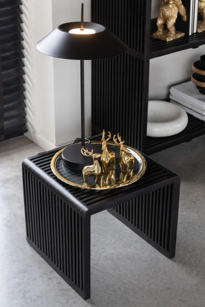 De Set van 2 salontafels Kubus Recycle Teak Zwart toont een gouden dienblad met drie gouden hertenfiguurtjes en een zwarte lamp. Daarachter staat een zwart rek met verschillende decoratieve accessoires.