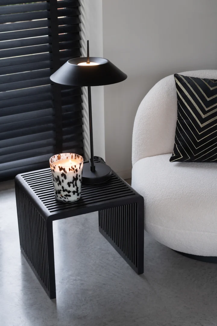 Een moderne set van twee Kubus Recycle Teak Black bijzettafels, gestyled met een zwarte lamp en een gevlekte kaars, staat naast een witte gebogen fauteuil met een kussen met goudpatroon. Zwarte jaloezieën bedekken het raam erachter.