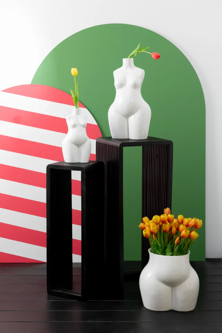 Twee witte vazen in de vorm van menselijke torso's en heupen met gele en oranje tulpen staan op een Set van 2 bijzettafels Kubus Recycle Teak Zwart, tegen kleurrijke gestreepte en groene abstracte achtergronden.
