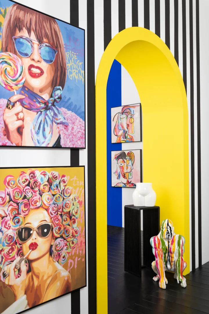 Een levendige kamer met zwart-wit gestreepte muren, kleurrijke pop-artportretten, een gewaagde gele boog en de Bijzettafel Zwart, een vierkante teakhouten bijzettafel van 34 x 34 cm, tentoongesteld naast een hurkende sculptuur met regenboogstrepen.