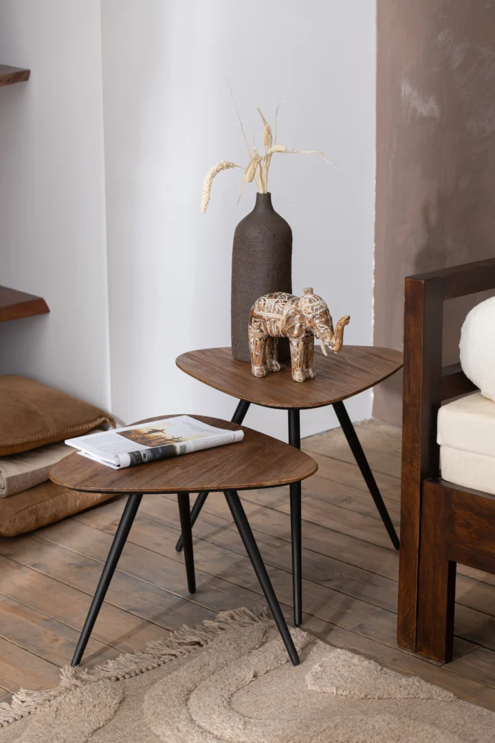 De Set van 2 tafels Driehoek Recycle Teak Natuurlijk bestaat uit twee houten bijzettafels met zwarte poten - de ene gedecoreerd met een donkere vaas met gedroogd gras en een olifant, de andere met een tijdschrift. Geplaatst naast een bank op een houten vloer met een beige vloerkleed.