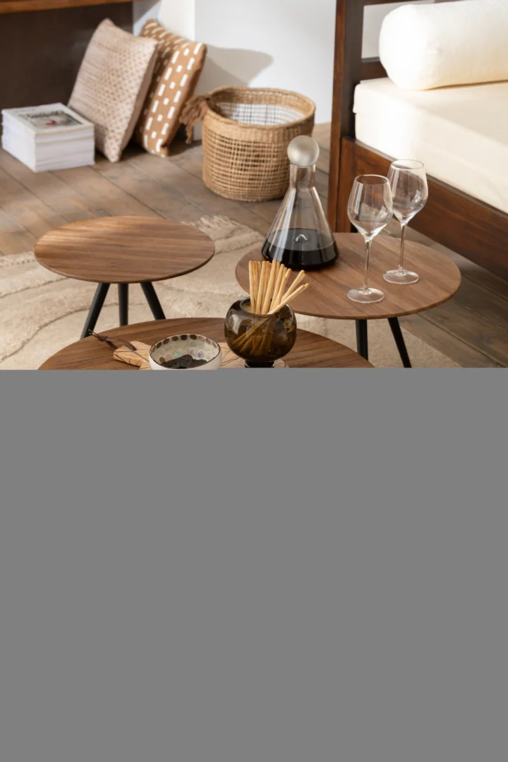 Een set van 4 ronde tafels van gerecycled teakhout natuurlijk met een karaf, twee wijnglazen, snacks en een vaas. Op de achtergrond een bank, kussens, boeken, vloerkleed en een mand voor een gezellige moderne woonkamer.