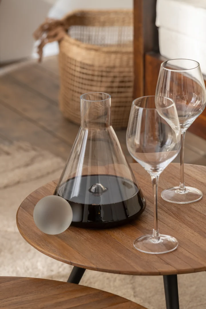 Een set van 4 ronde tafels van gerecycled teakhout natuurlijk is te zien, met daarop een glazen wijnkaraf gevuld met rode wijn en twee lege wijnglazen. Op de achtergrond staan een rieten mand en een deel van een bedframe.